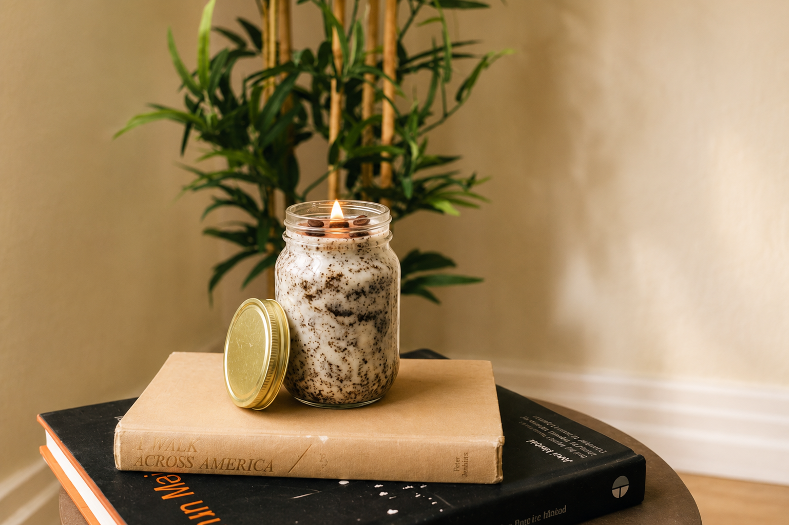 A lit candle in a glass jar studded with coffee beans, its gold lid resting beside it on a stack of books.